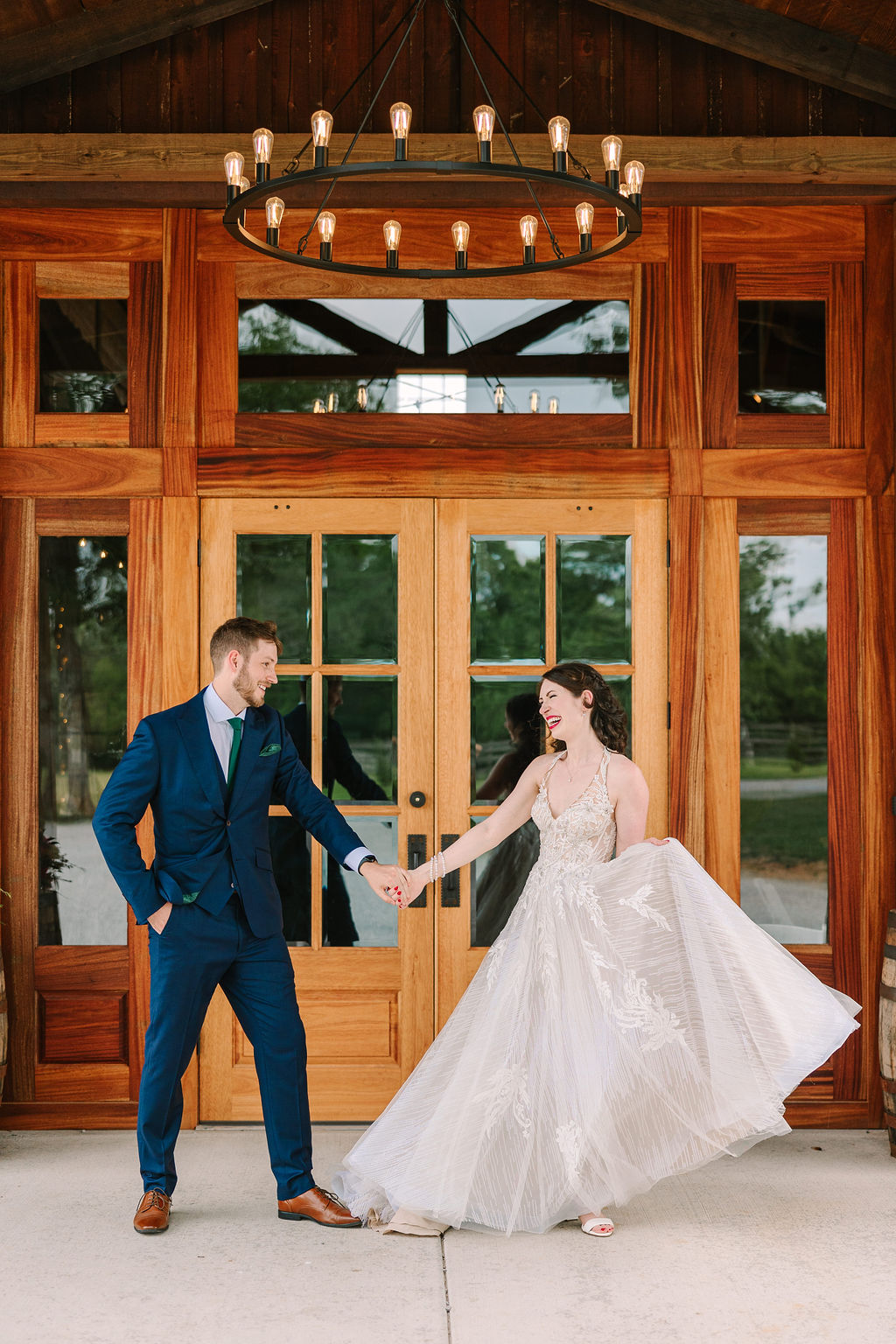 Kate & Josh laugh hand-in-hand under Covey Creek’s barn porch chandelier, her tulle skirt swirling