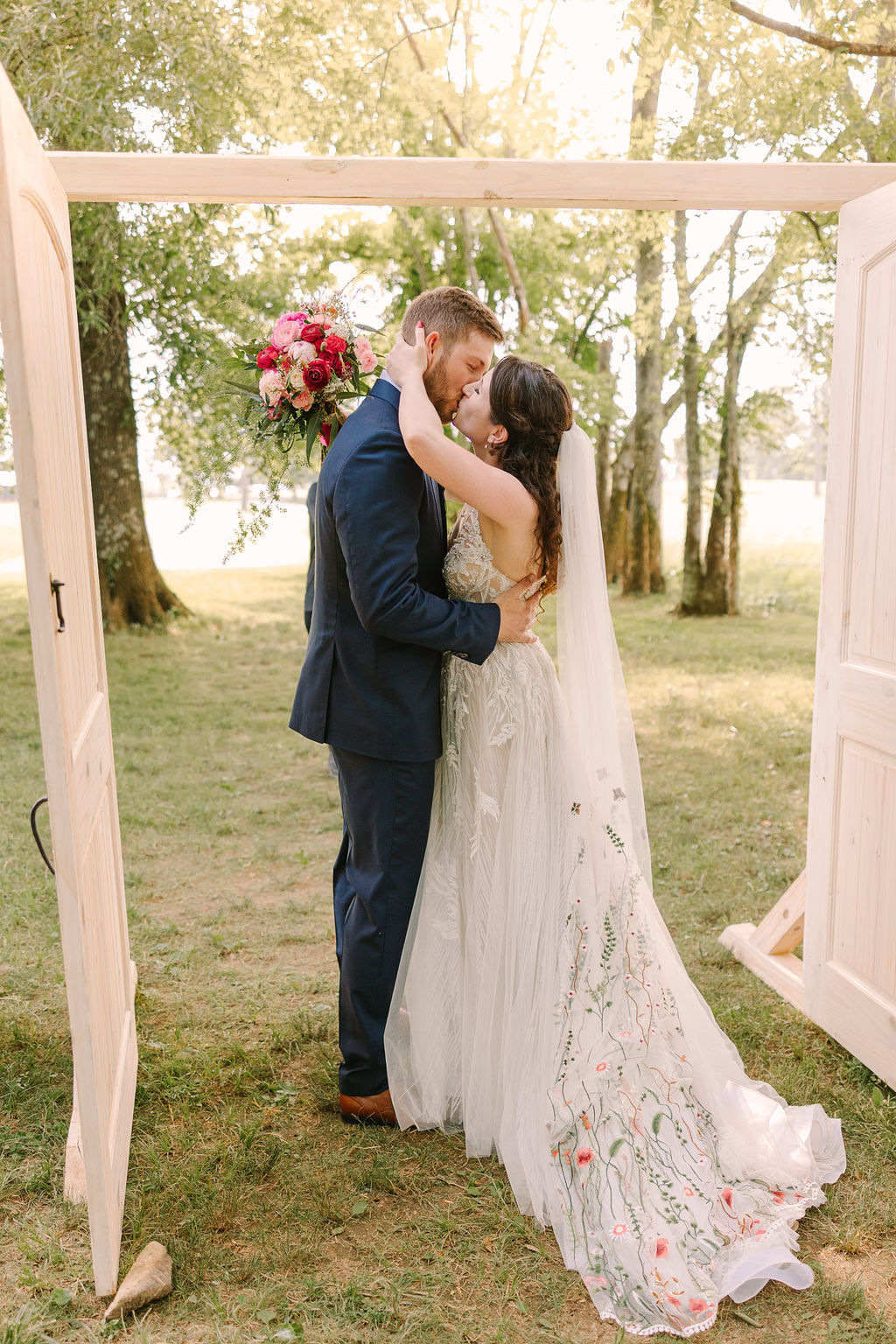 Kate & Josh kiss at Covey Creek, framed by open wooden doors, her floral-embroidered veil trailing