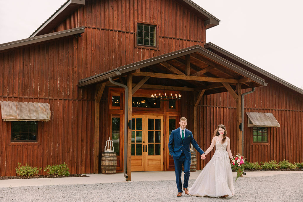 Kate & Josh walk hand-in-hand past Covey Creek’s barn entrance, pink bouquet and green tie in view