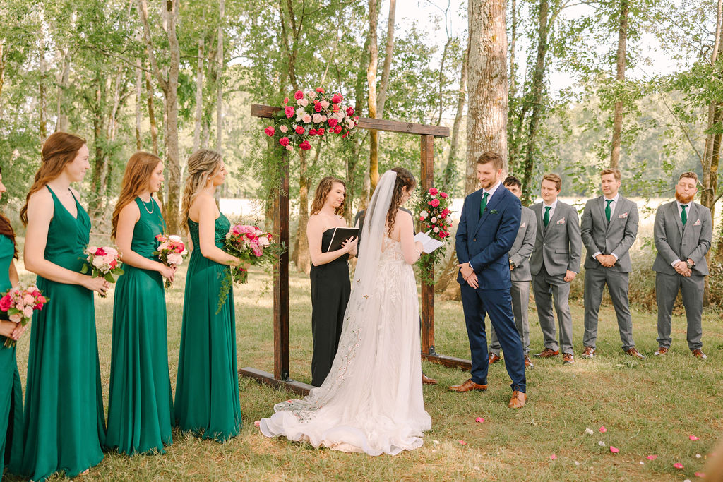 Kate & Josh exchange vows at Covey Creek under a floral wooden arch, bridal party in emerald and grey