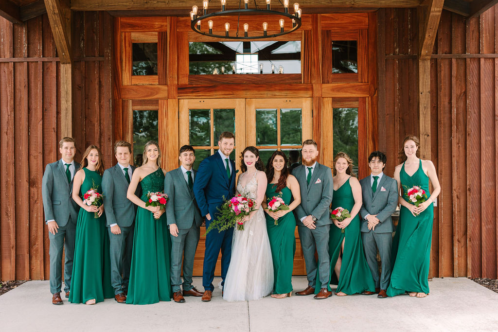 Kate & Josh pose with their bridal party in emerald and grey under Covey Creek’s barn porch chandelier