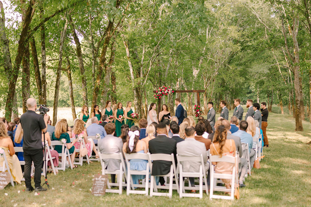 Wide view of Kate & Josh’s Covey Creek ceremony under a floral arch among trees, guests in white chairs