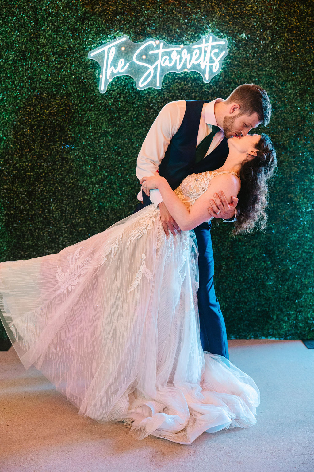 Kate & Josh share a dip kiss before Covey Creek’s greenery wall and The Starretts neon sign
