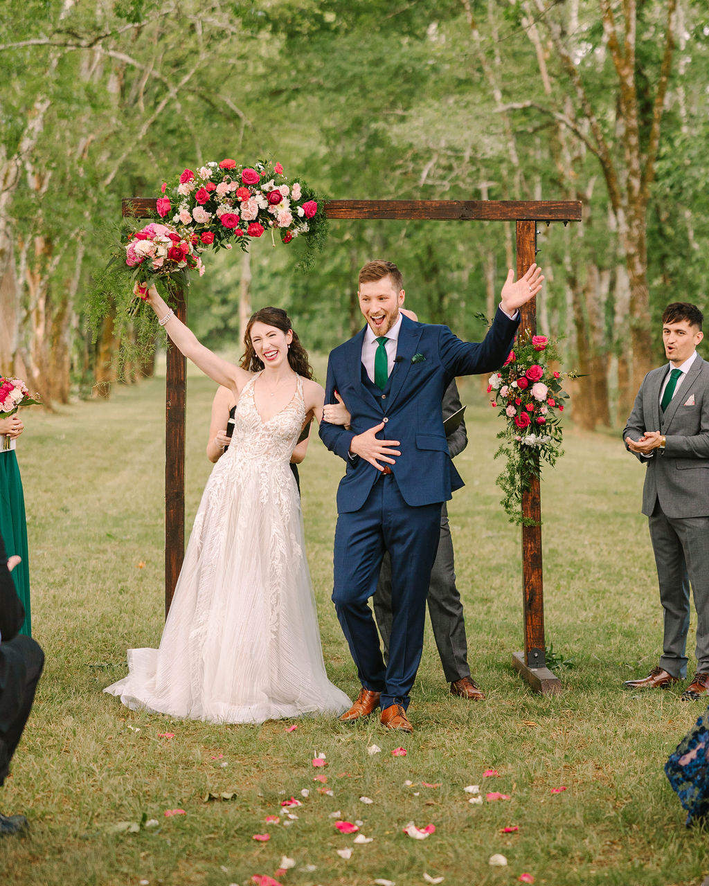 Kate & Josh cheer down the aisle at Covey Creek, pink bouquet raised past a floral wooden arch