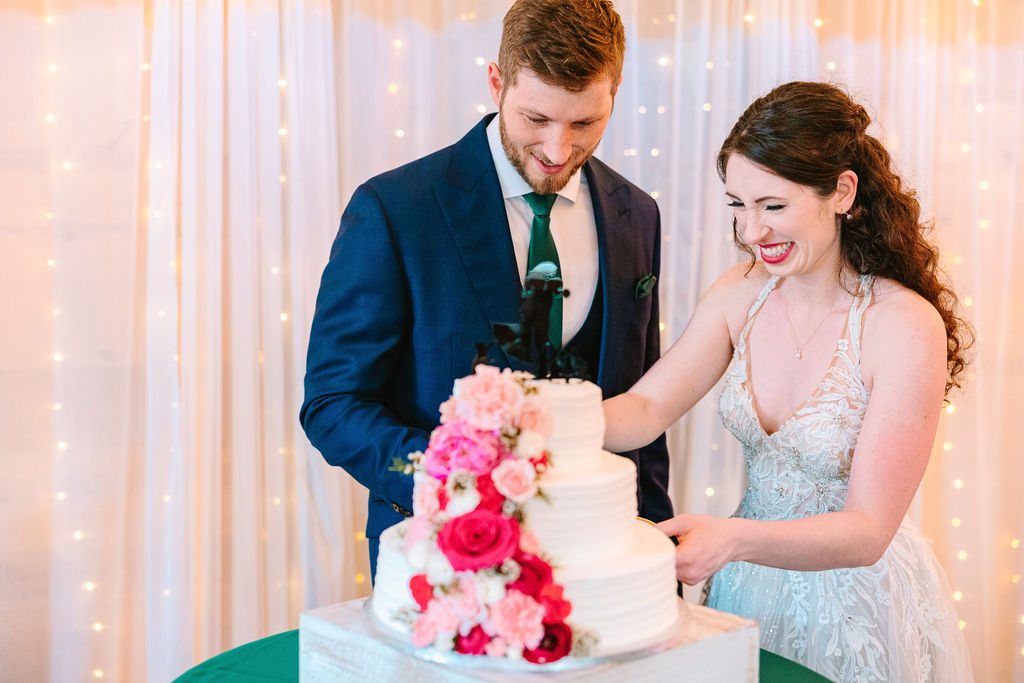 Kate & Josh laugh while cutting their three-tier cake with pink roses at Covey Creek’s reception