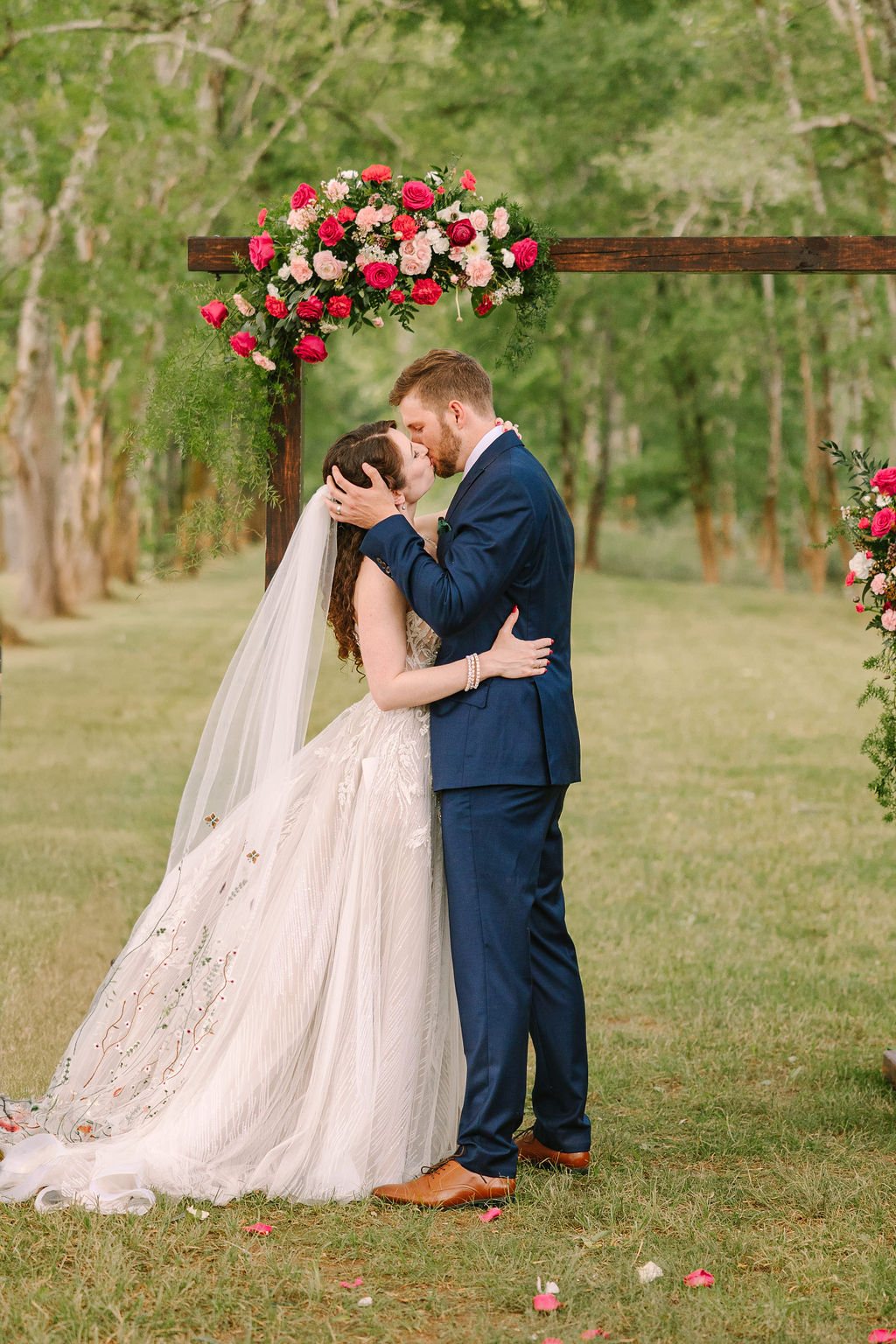 Kate & Josh kiss under Covey Creek’s floral wooden ceremony arch on a petal-strewn grassy aisle