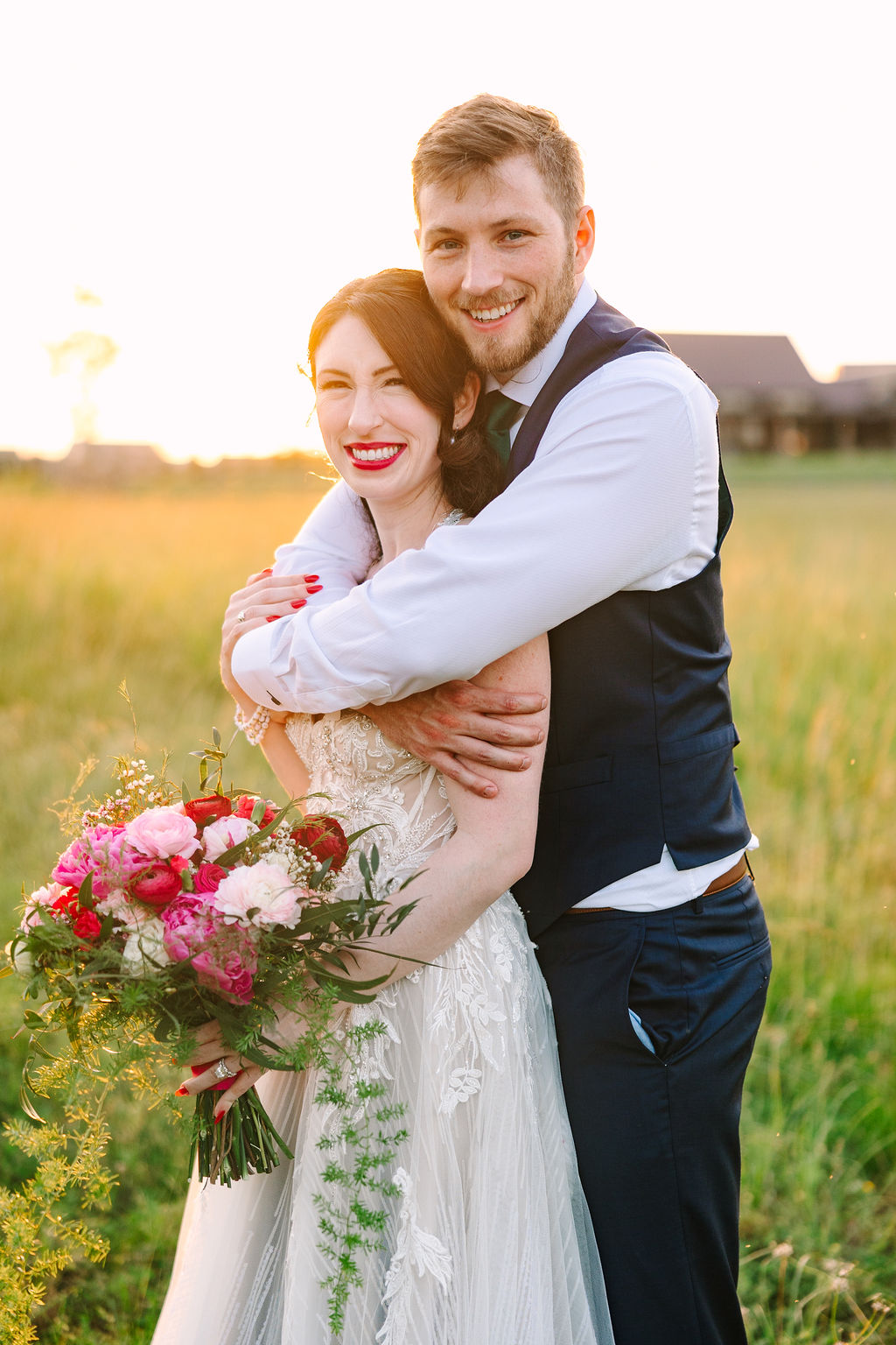 Kate & Josh hug and smile at golden hour in Covey Creek’s tall grass, pink-red bouquet against navy vest