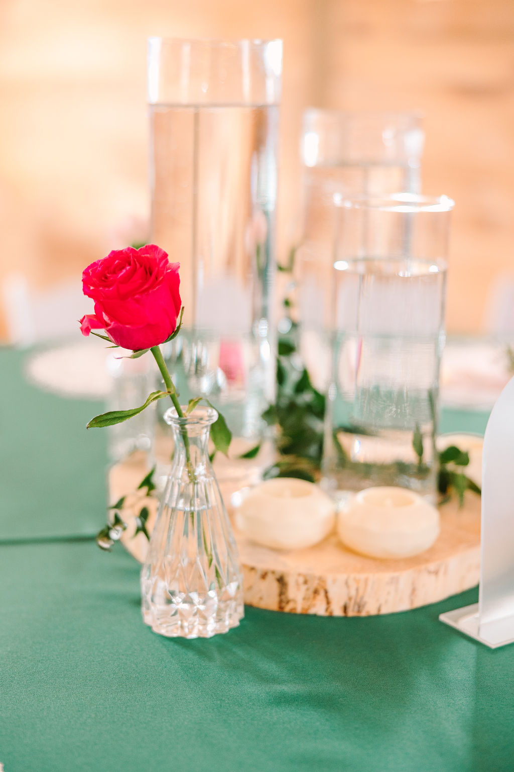 Kate & Josh Covey Creek detail: pink rose in a bud vase beside glass cylinders on a wood-slice centerpiece
