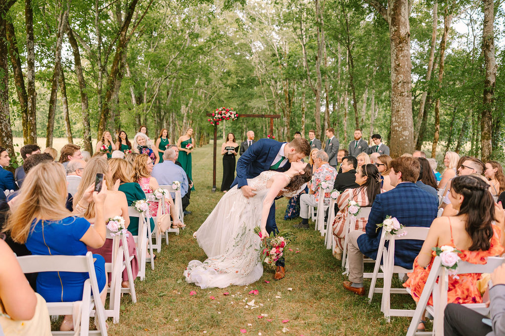 Kate & Josh share a dip kiss in Covey Creek’s wooded aisle as guests cheer from white folding chairs