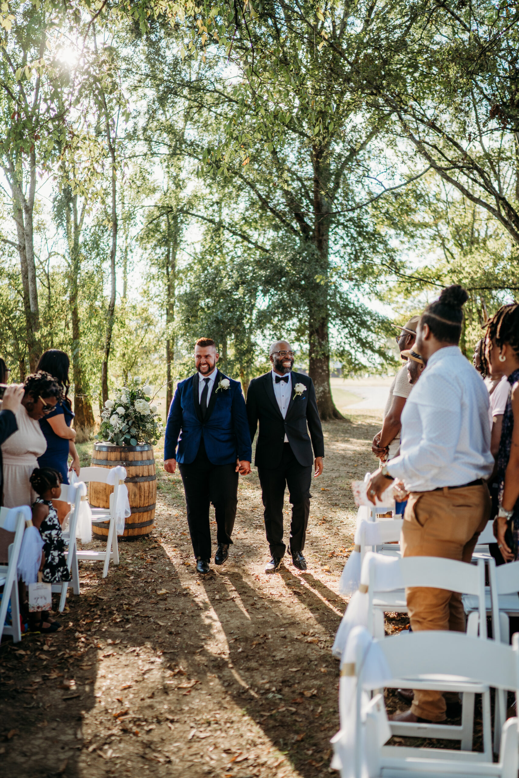 Levi & Billy smile walking hand-in-hand down the Tunnel of Love aisle past white chairs at Covey Creek