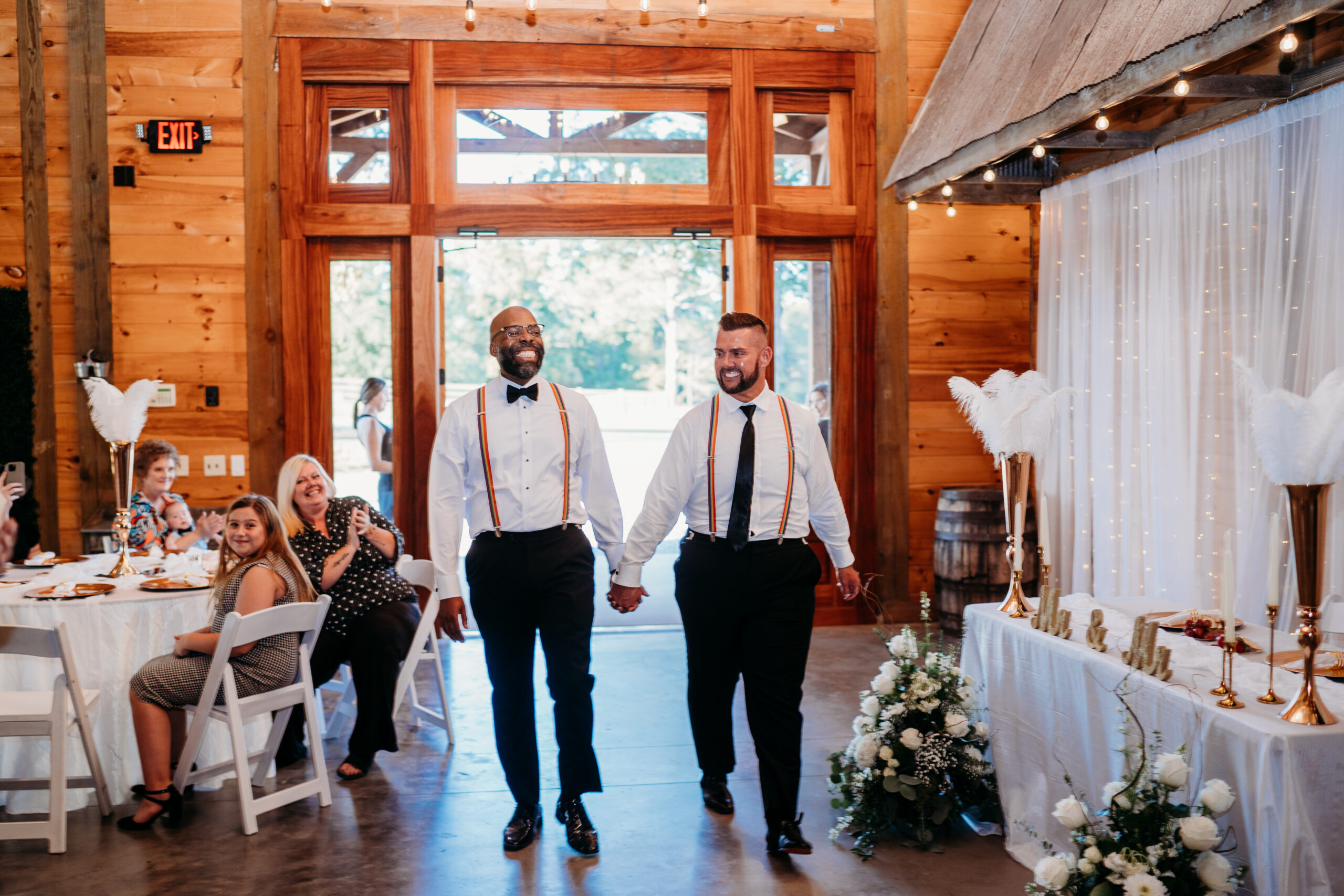 Levi & Billy walk laughing past Mr & Mr feather head table in rainbow suspenders at Covey Creek