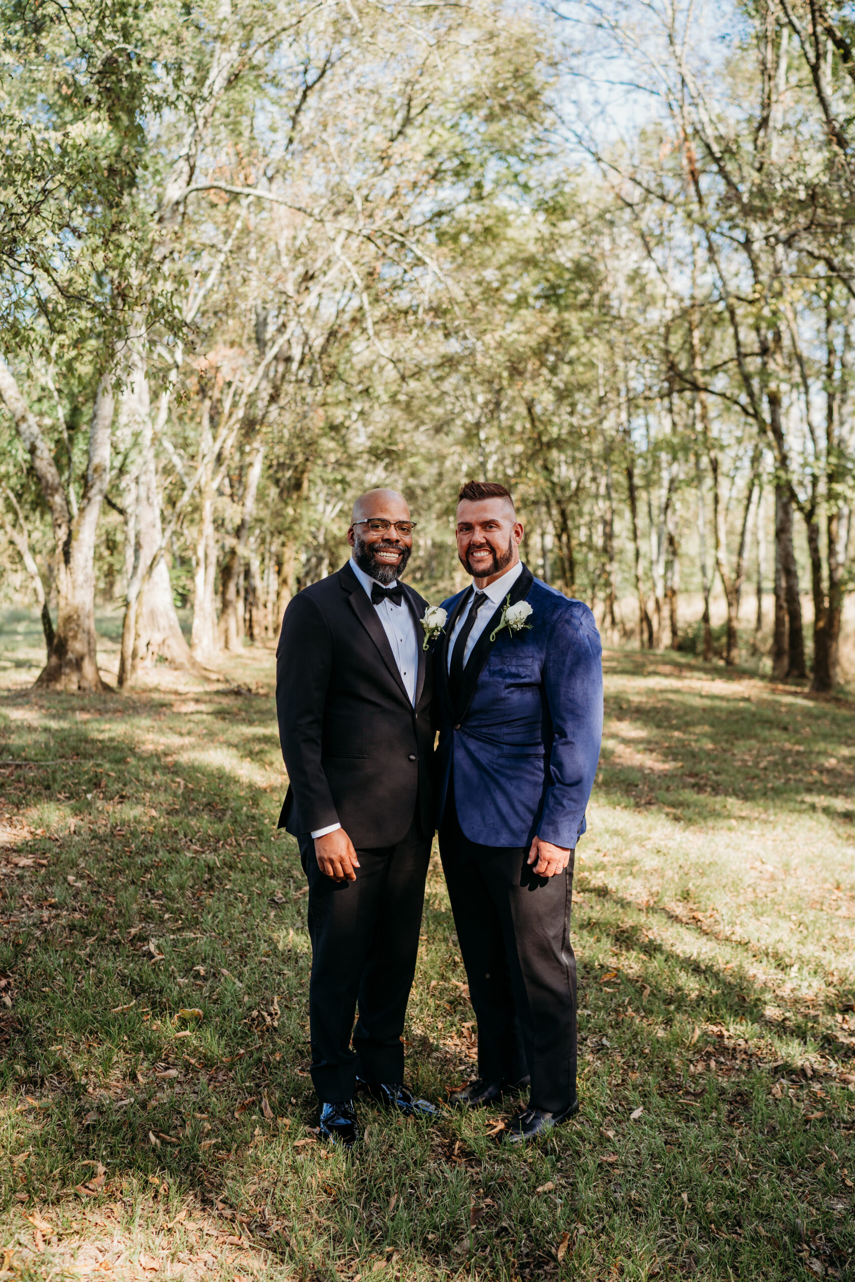 Levi & Billy smile side-by-side in black tuxedo and blue velvet jacket among Covey Creek trees