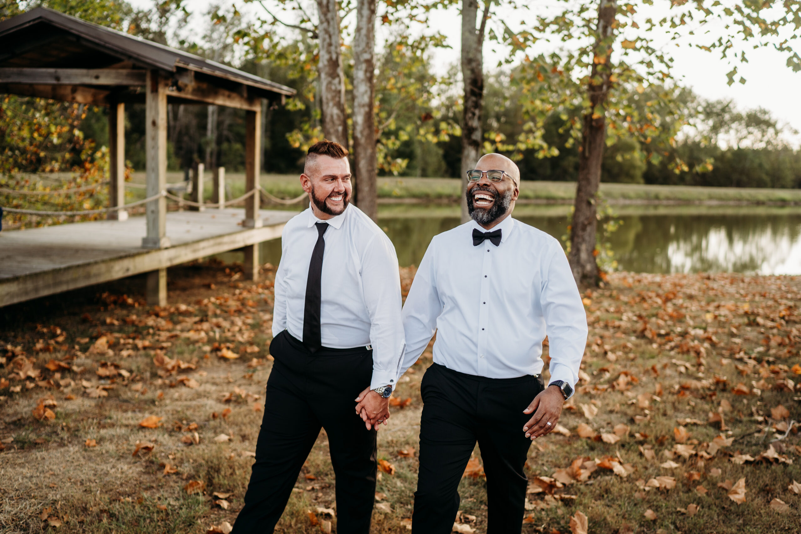 Levi & Billy laugh walking hand-in-hand past Covey Creek Pond dock in white shirts and ties