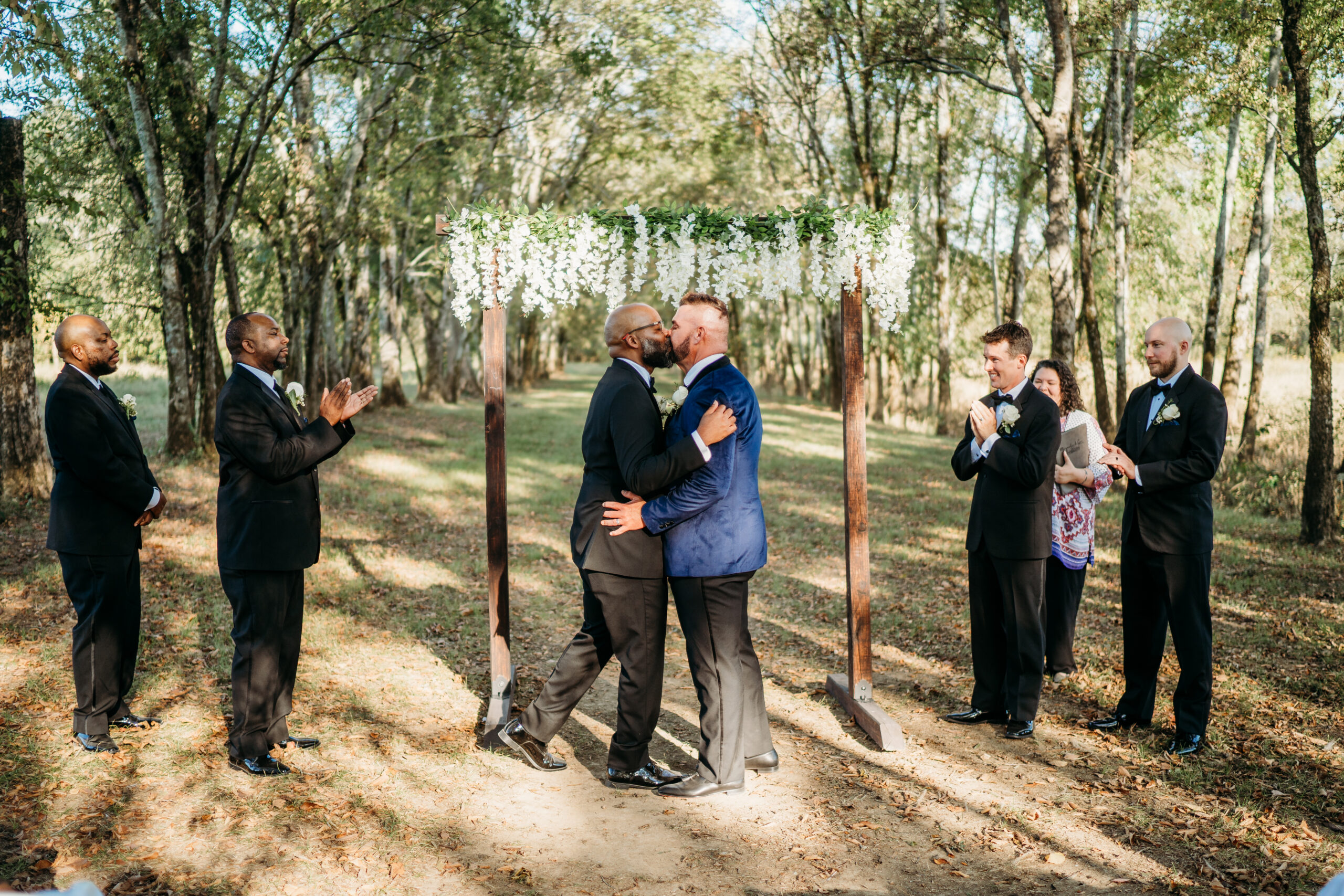 Levi & Billy kiss under white-flowered arbor as groomsmen clap on Covey Creek Tunnel of Love