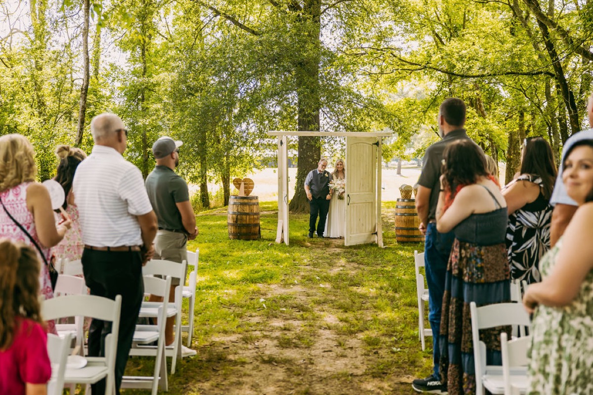Alexis & Logan: Alexis enters through open white doors down the wooded aisle as guests rise at Covey Creek