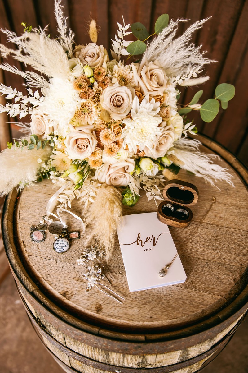 Alexis & Logan bridal flat lay on a barrel: cream bouquet, her vows book, rings, and hairpins at Covey Creek