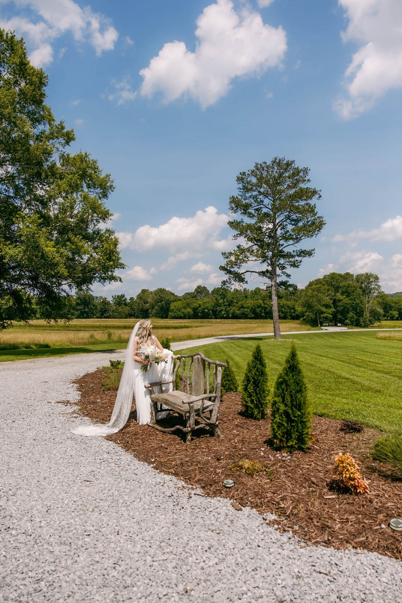Alexis & Logan: Alexis stands by a rustic branch bench on the open lawn under blue sky at Covey Creek