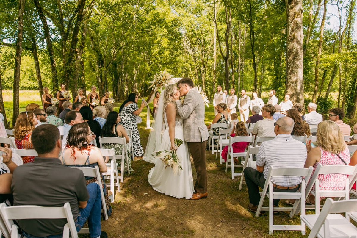 Alexis & Logan kiss at the altar as guests watch from white chairs under the trees at Covey Creek