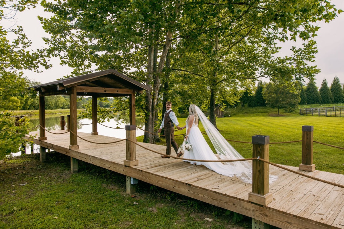Alexis & Logan stroll the Pond pavilion dock hand in hand with her long veil trailing at Covey Creek