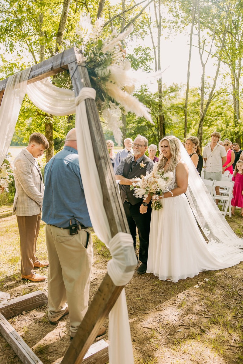 Alexis & Logan under the pampas-draped wood arch as Alexis smiles during the ceremony at Covey Creek
