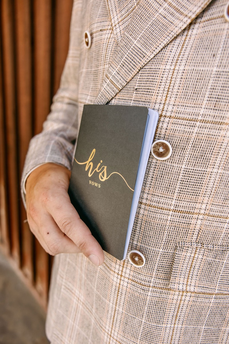 Alexis & Logan: Logan holds his vows book against a tan plaid double-breasted jacket at Covey Creek