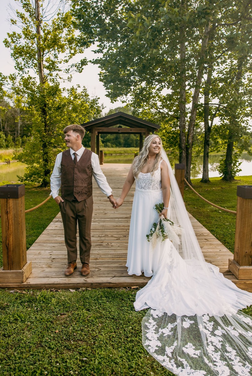 Alexis & Logan stand hand in hand on the Pond dock with veil trailing and gazebo behind at Covey Creek