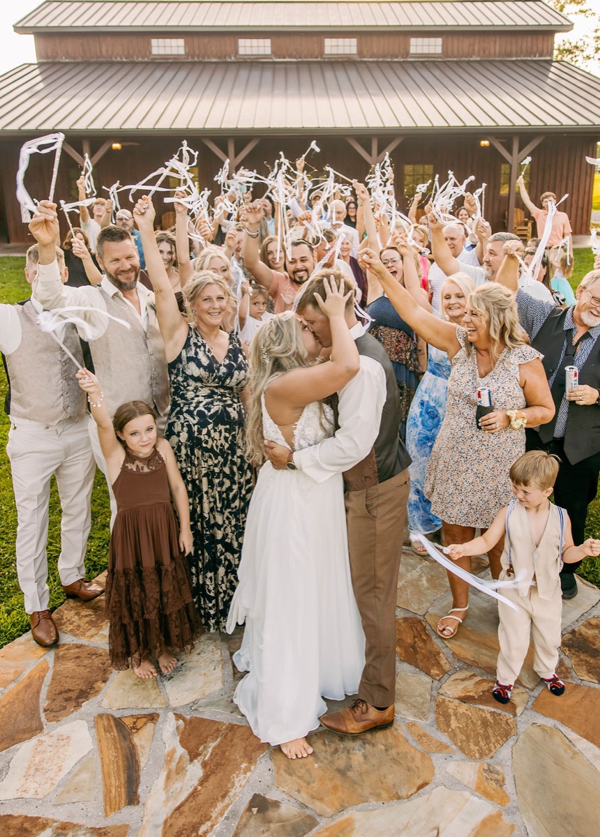Alexis & Logan kiss on the patio as guests cheer with ribbon wands outside the barn at Covey Creek