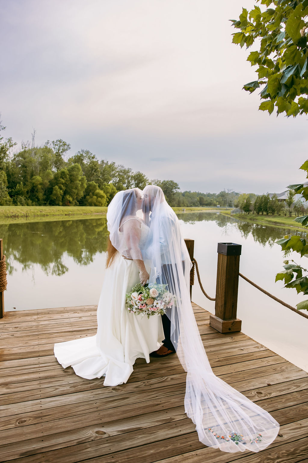 Amanda & Stephen kiss under a shared veil on Covey Creek’s Pond dock, embroidered train on the planks