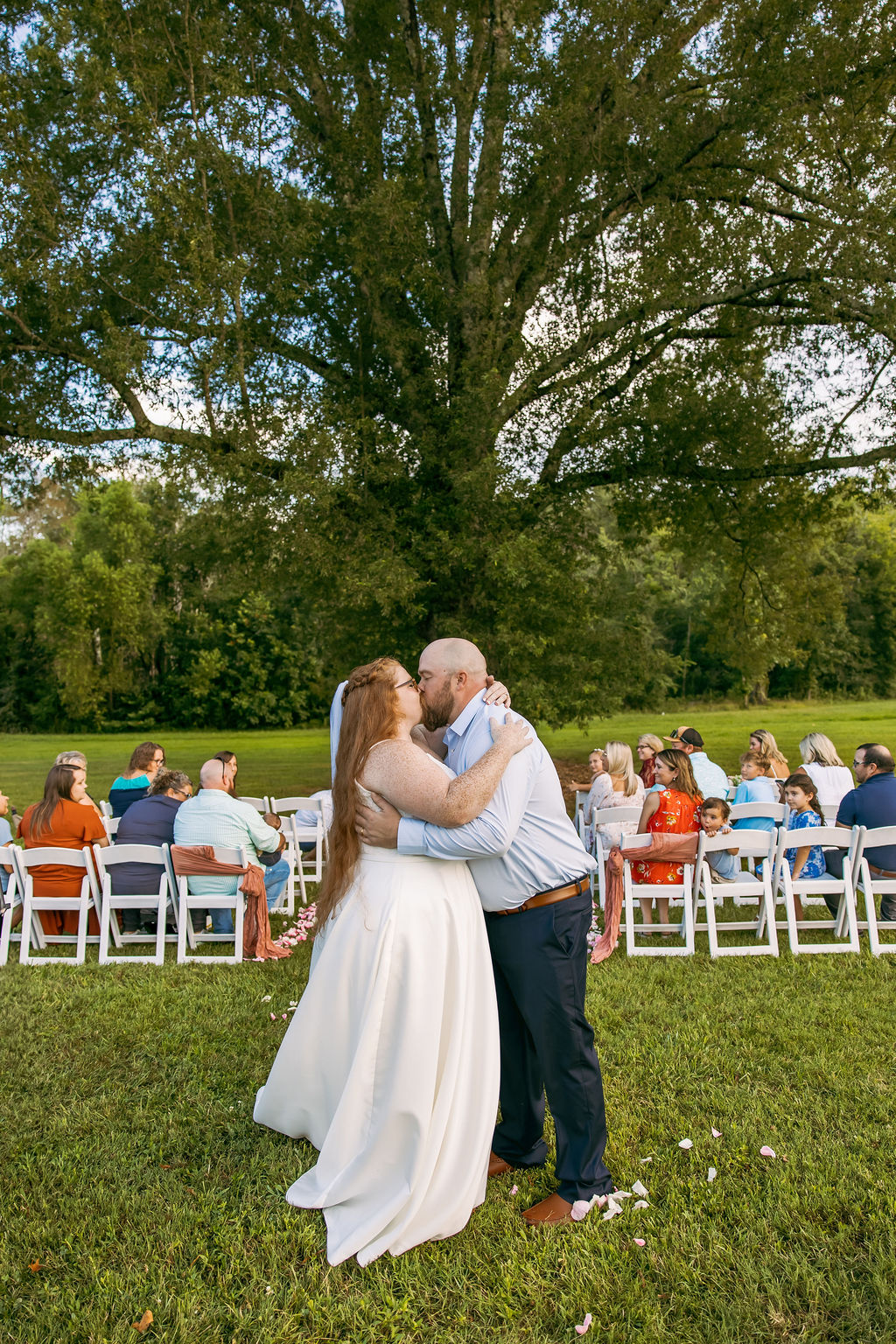 Amanda & Stephen kiss under Covey Creek’s Tunnel of Love canopy with petal-strewn grass and seated guests