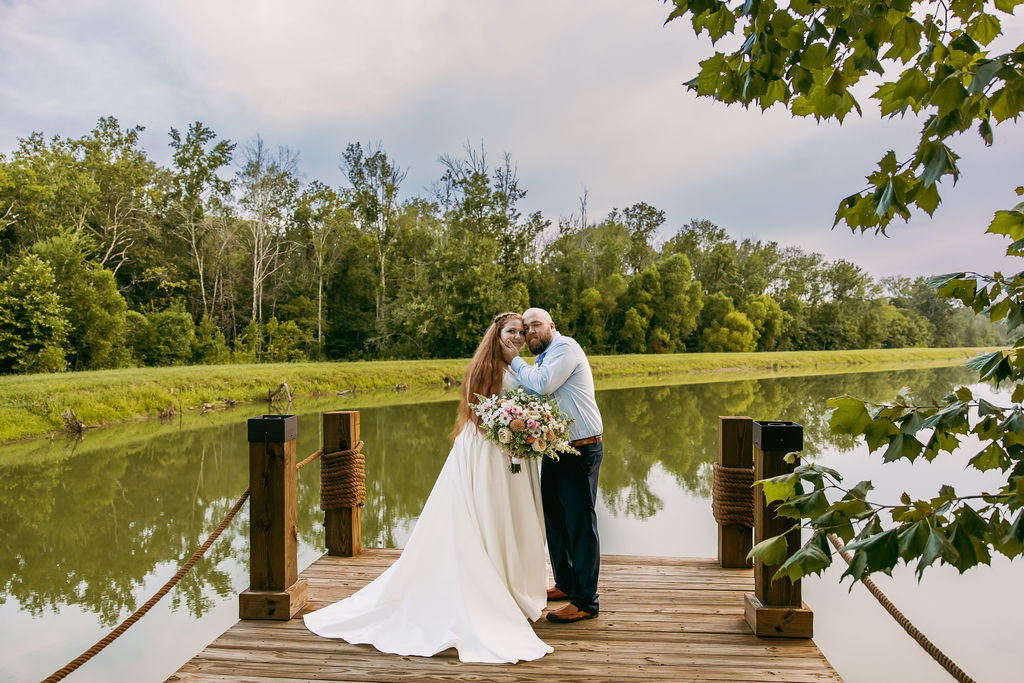 Amanda & Stephen pose on Covey Creek’s Pond dock as he cups her cheek, rope-wrapped posts beside them