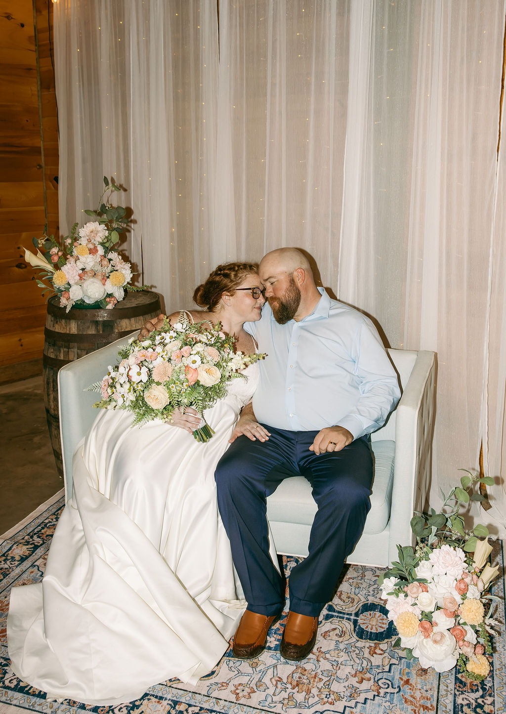 Amanda & Stephen sit forehead-to-forehead on a Covey Creek lounge sofa with bouquet and barrel florals