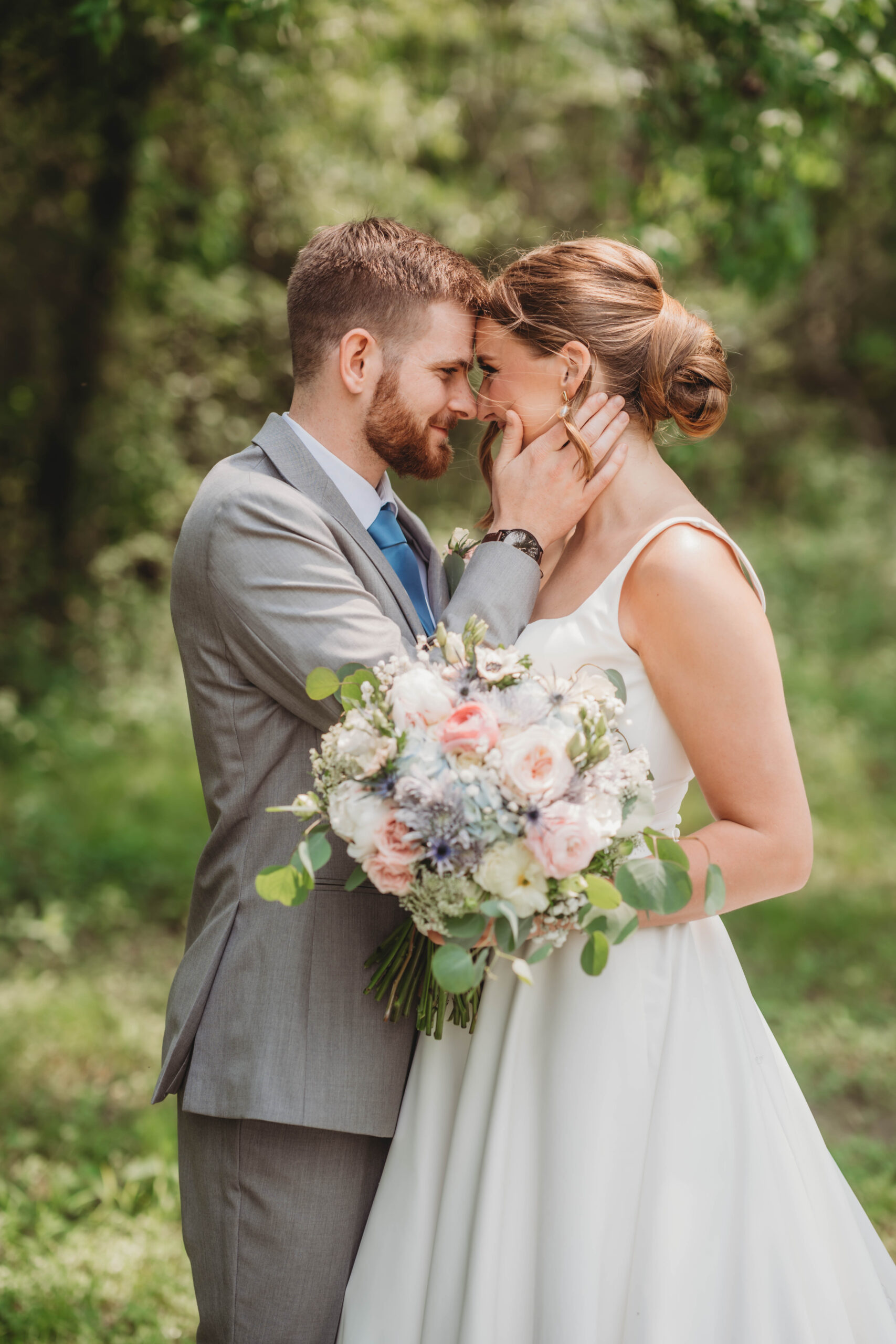 Anna & Jaden press foreheads together outdoors, her pink and blue bouquet in hand, at Covey Creek.