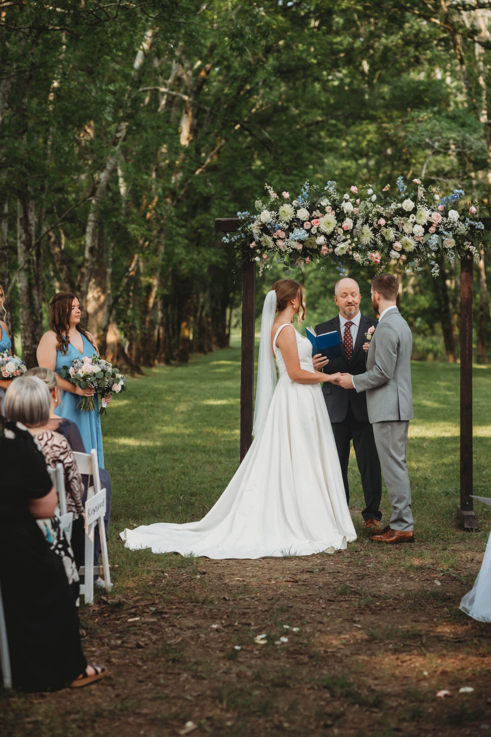 Anna & Jaden hold hands under a floral wooden arch as their officiant reads vows at Covey Creek.