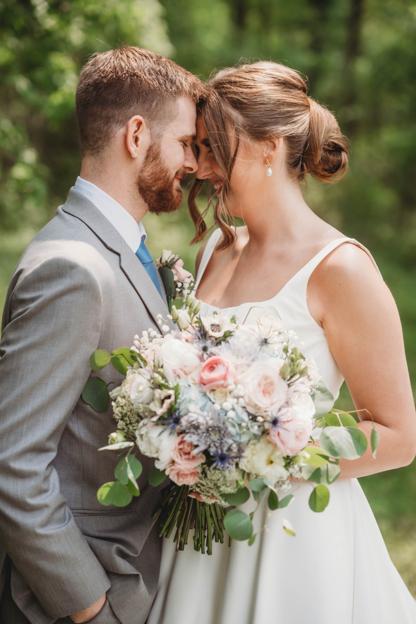 Anna & Jaden touch foreheads, her pink rose and blue thistle bouquet between them, at Covey Creek.