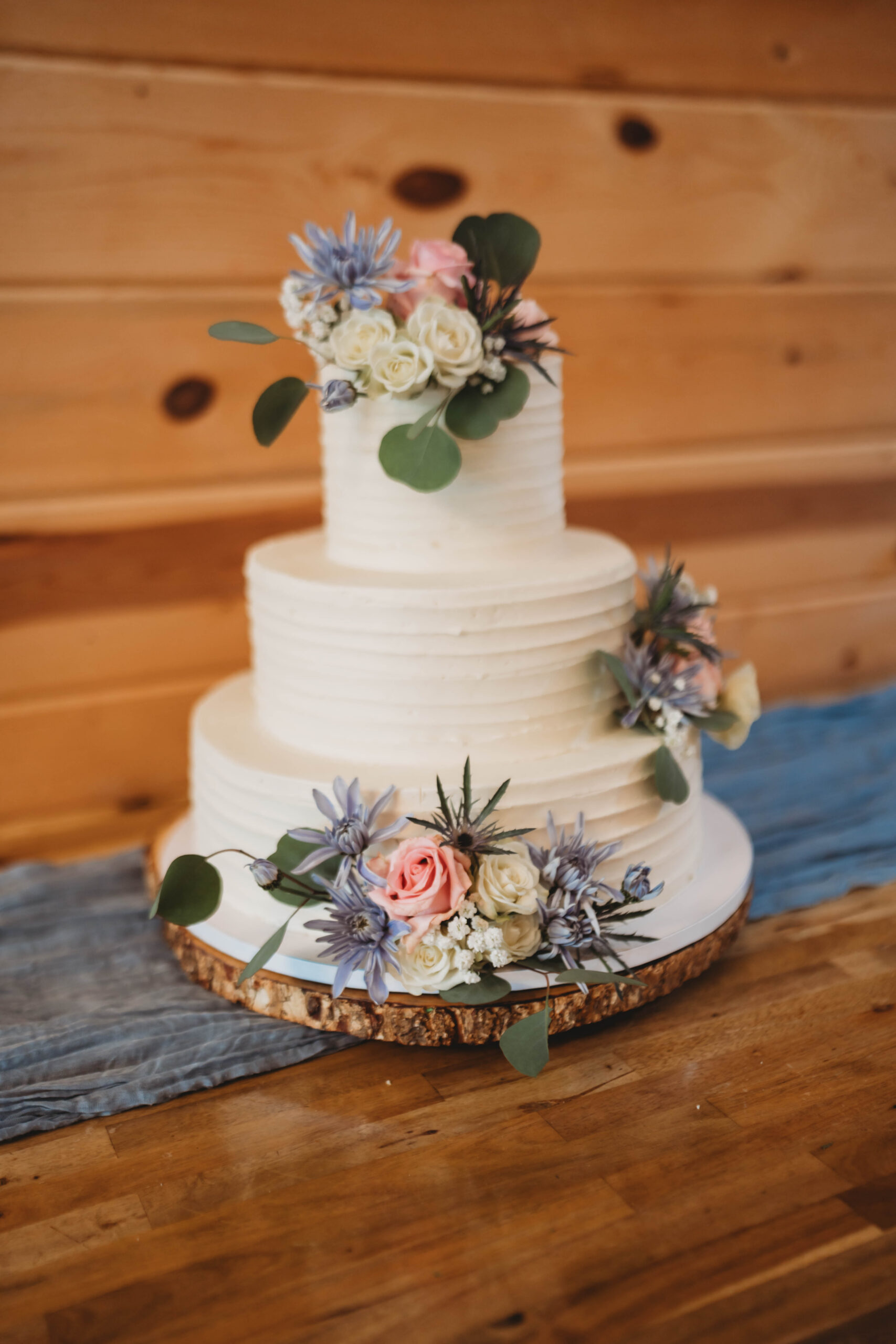 Three-tier white cake with pink roses and blue florals on a wood stand for Anna & Jaden at Covey Creek.