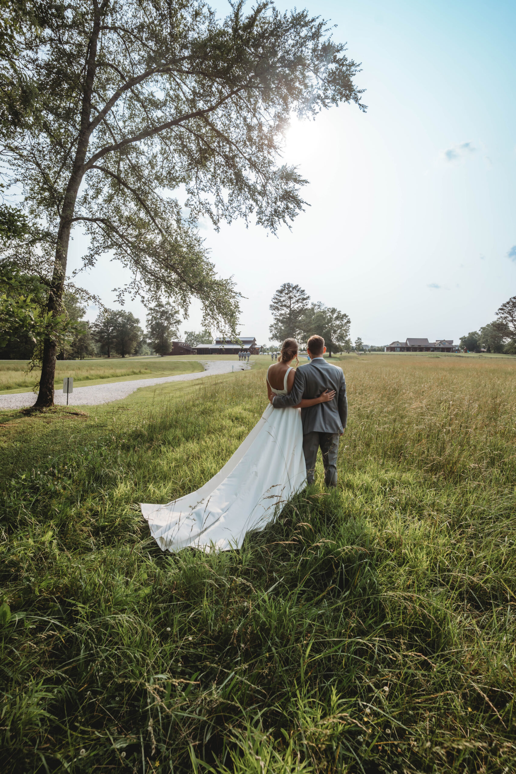 From behind, Anna & Jaden look across open fields toward the venue barns under blue sky at Covey Creek.
