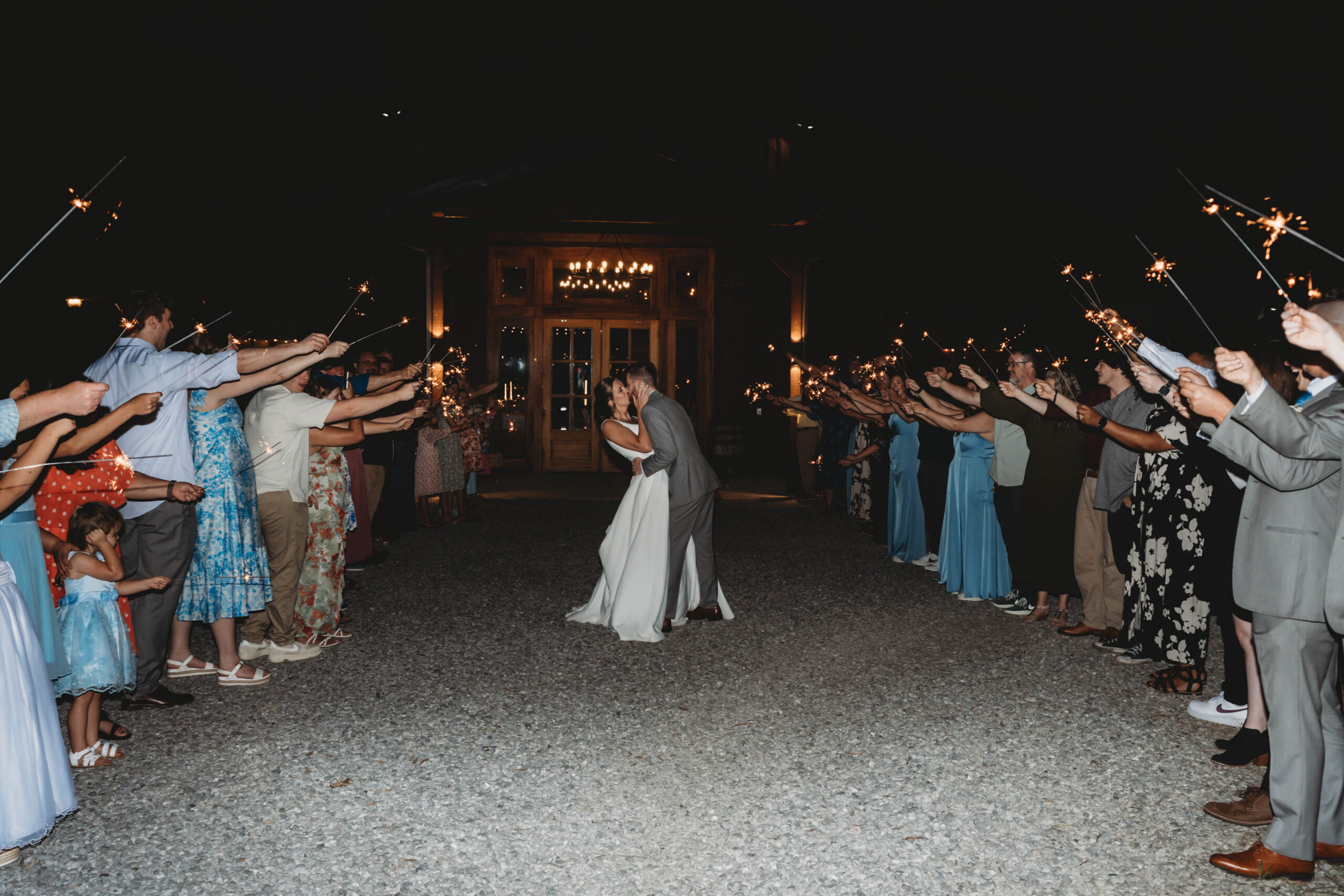 Anna & Jaden kiss in a sparkler tunnel as guests line the gravel path outside at Covey Creek.