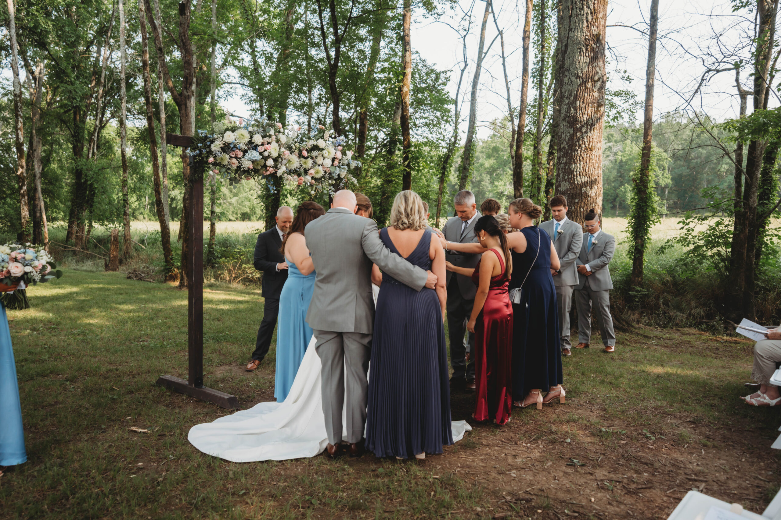 Anna & Jaden gather with family in a prayer circle under the floral ceremony arch at Covey Creek.
