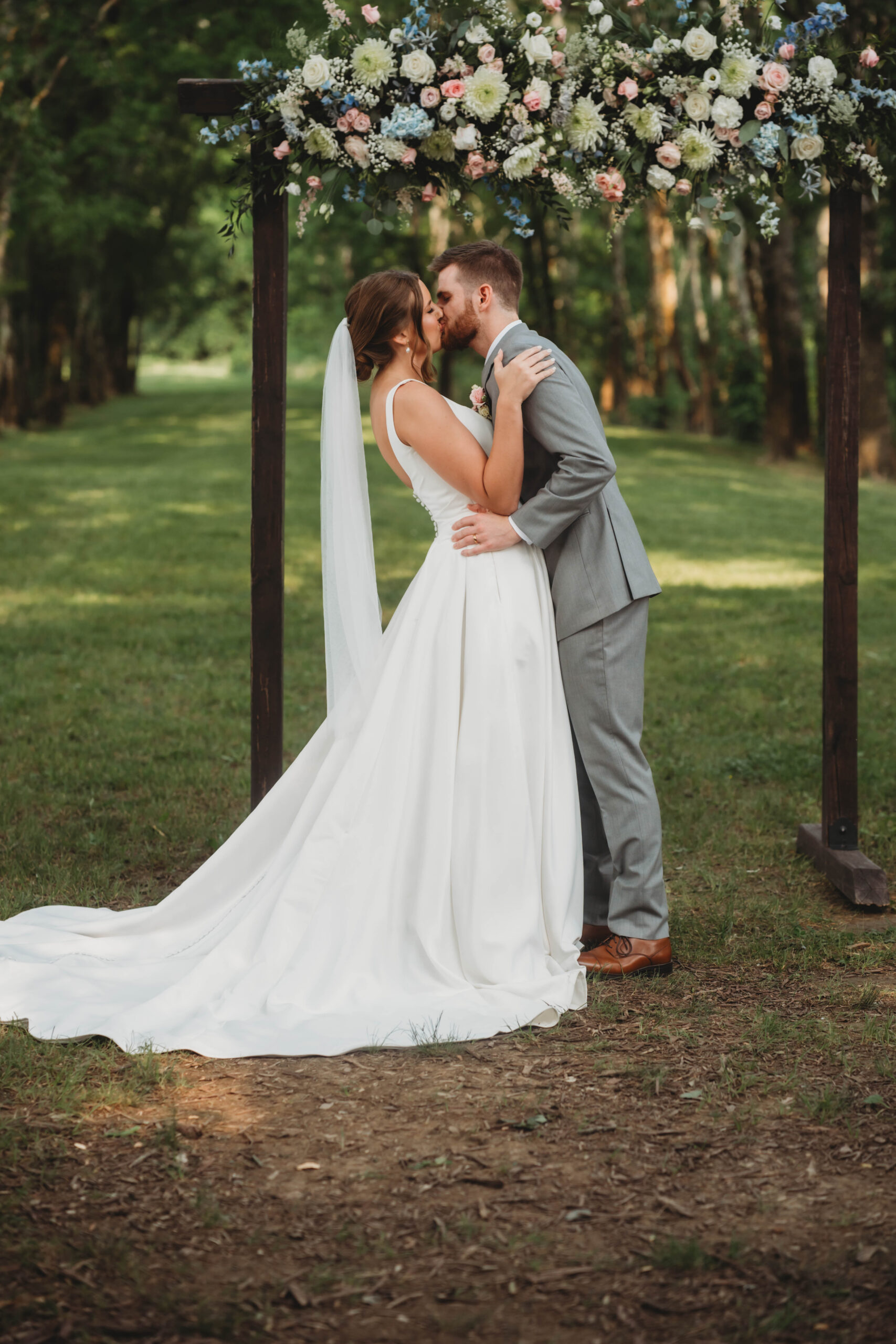 Anna & Jaden share their first kiss under a floral-topped wooden arch outdoors at Covey Creek.