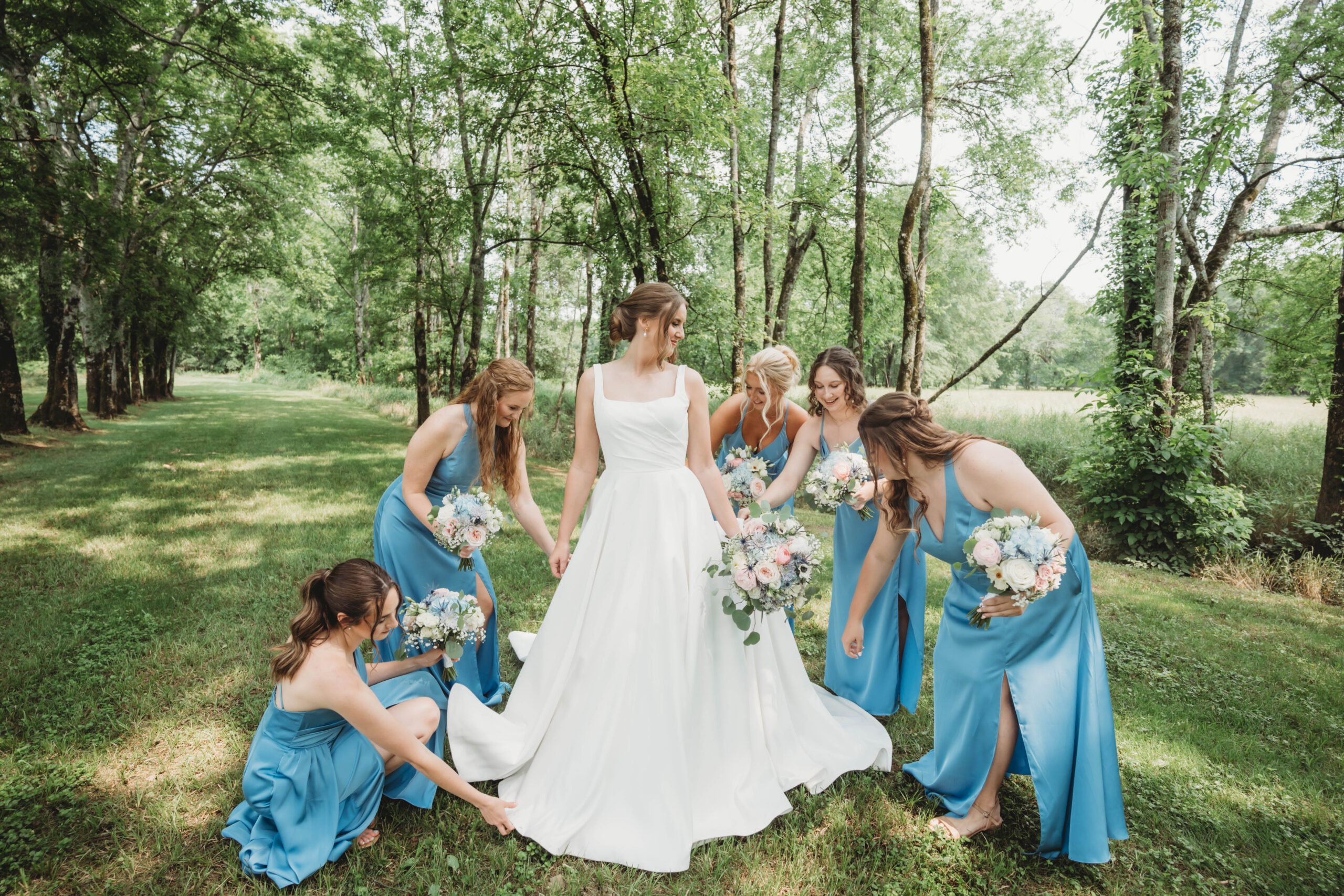 Bridesmaids in dusty blue fluff Anna's gown along a wooded path for Anna & Jaden at Covey Creek.