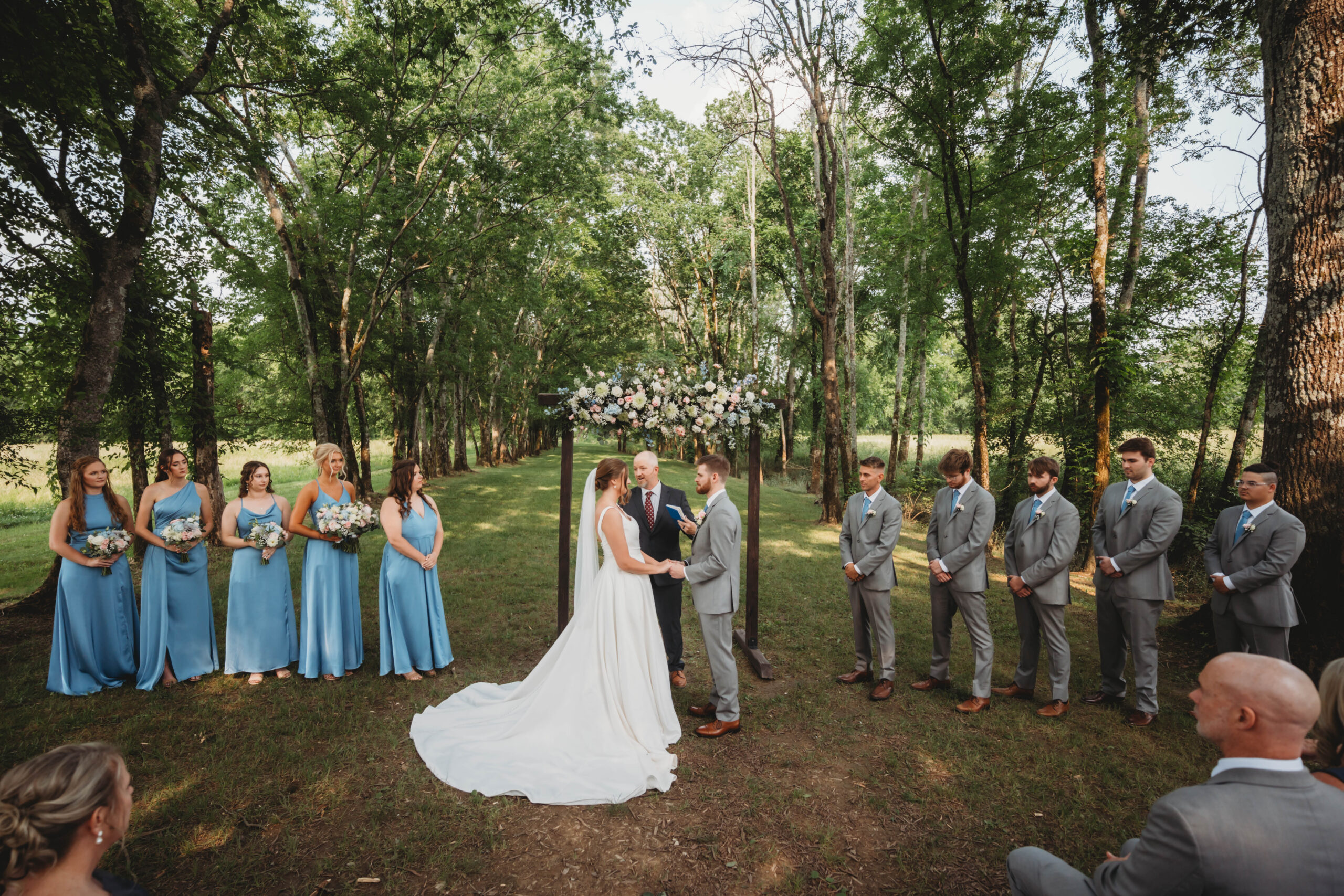 Anna & Jaden exchange vows under a floral wooden arch as their party and guests watch at Covey Creek.