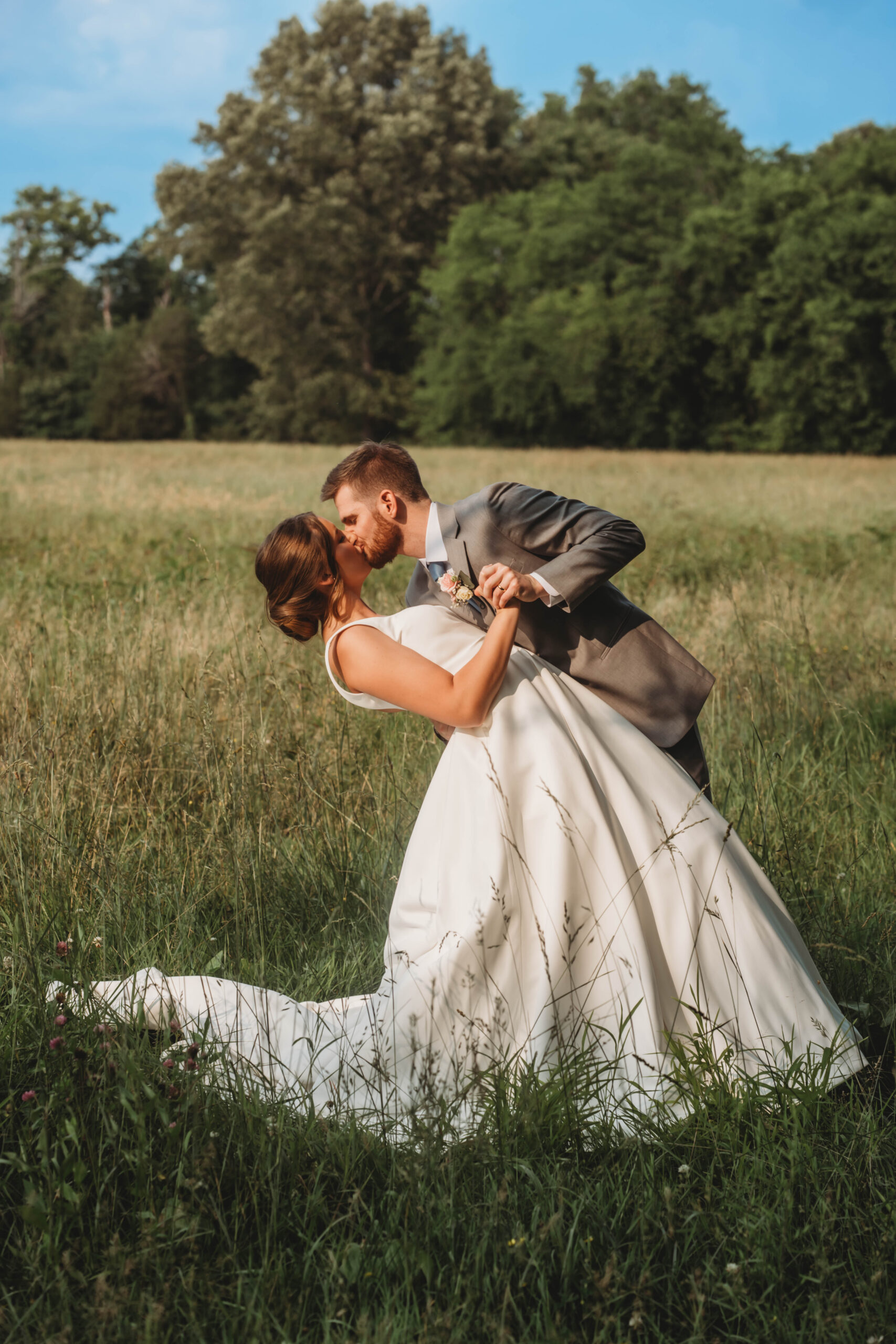 Anna & Jaden share a romantic dip kiss in a tall grassy field under blue sky at Covey Creek.