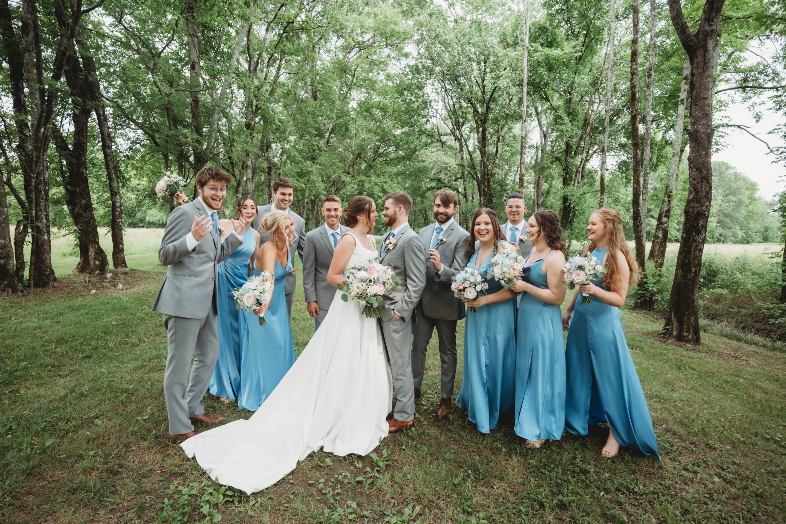 Anna & Jaden stand with their wedding party in dusty blue and grey suits among trees at Covey Creek.