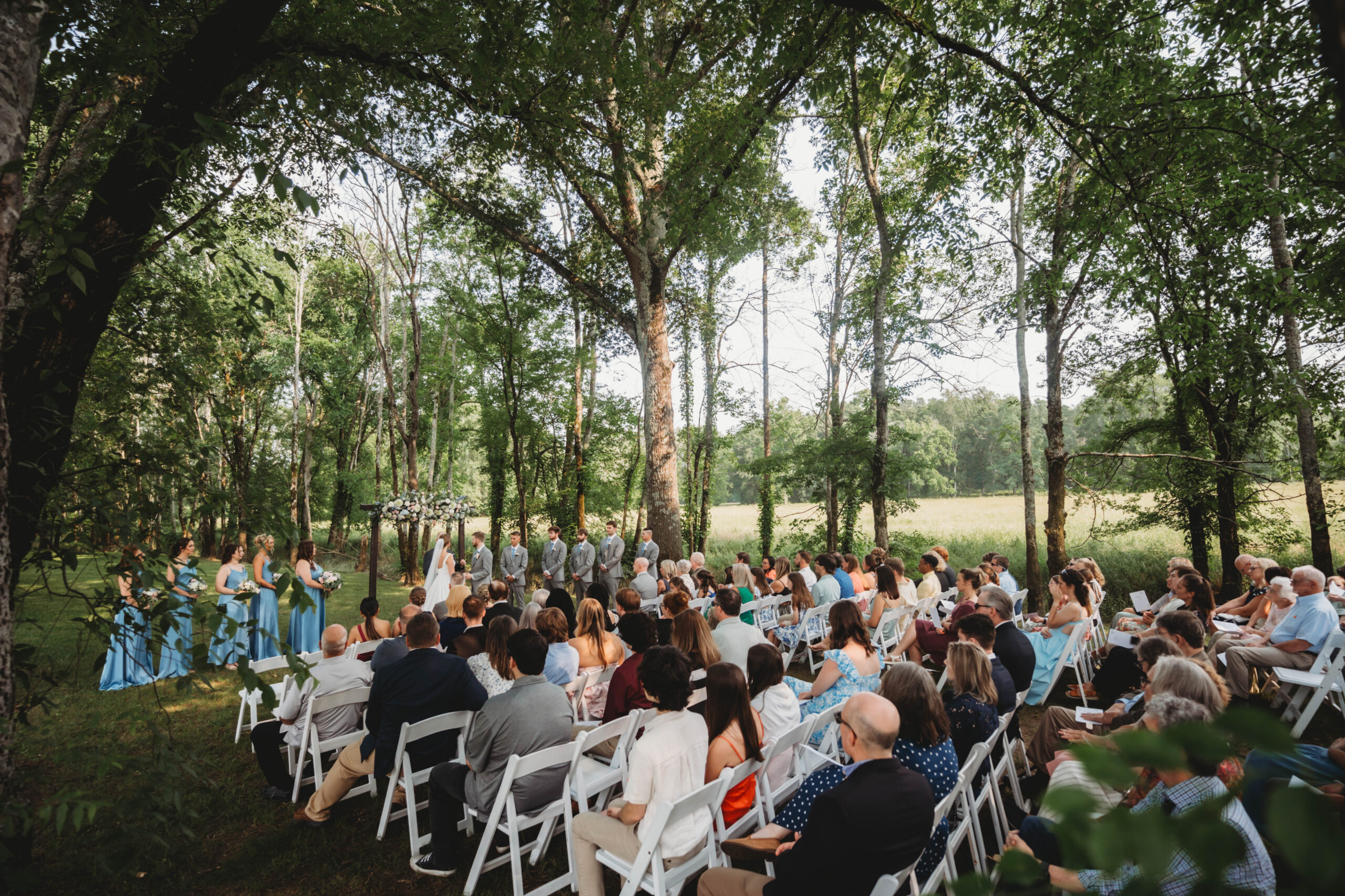 Anna & Jaden stand at the floral arch while guests watch from white chairs under trees at Covey Creek.