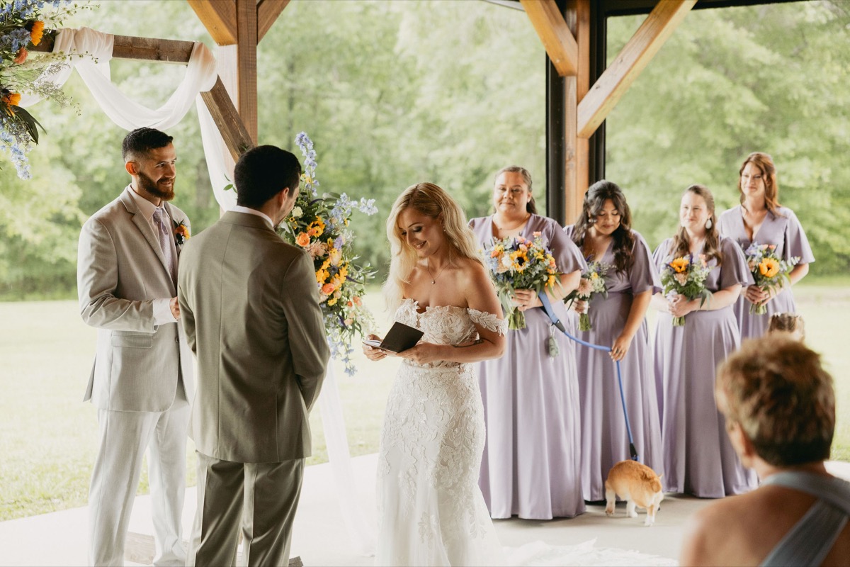 Brianna & Michael exchange vows as she reads a black book; lavender bridesmaids and a corgi stand at Covey Creek.