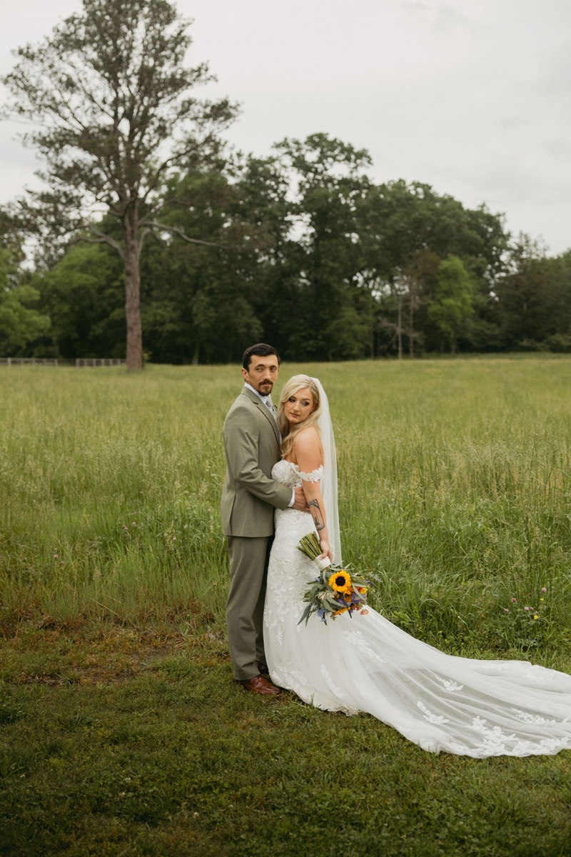Brianna & Michael pose in a tall grassy field, her lace train spread, sunflower bouquet in hand, at Covey Creek.