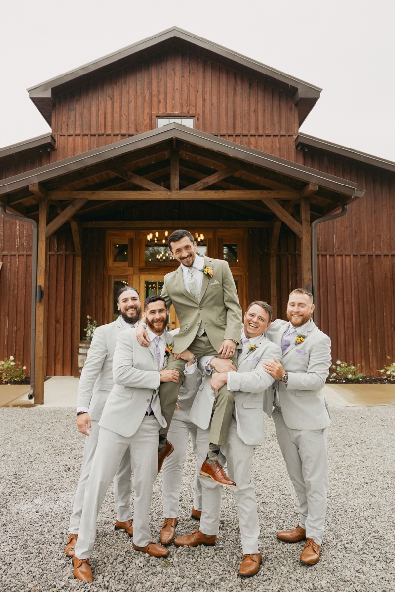 Groomsmen hoist Michael in a sage suit before Covey Creek's barn doors for Brianna & Michael's wedding day.