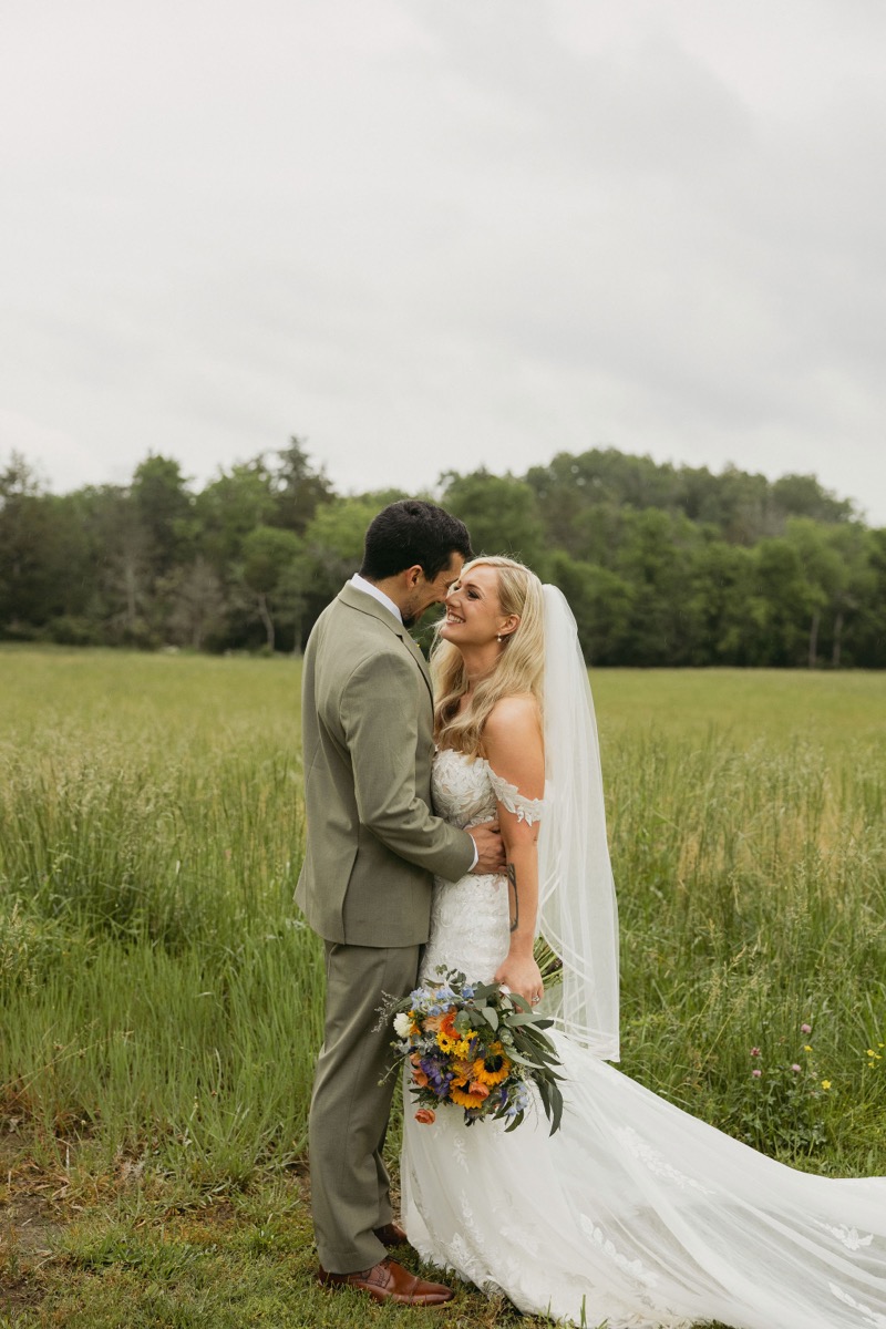Brianna & Michael face each other smiling in tall grass, sunflower bouquet and lace train visible, at Covey Creek.