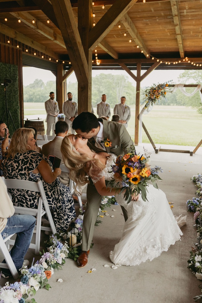Brianna & Michael share a dip kiss in the aisle, sunflower bouquet in hand, as guests cheer at Covey Creek's pavilion.