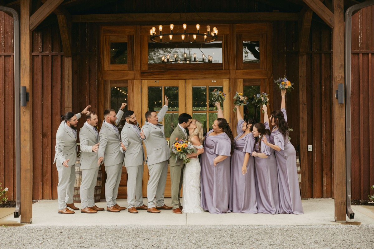 Wedding party cheers as Brianna & Michael kiss under a porch chandelier at the Covey Creek barn doors.